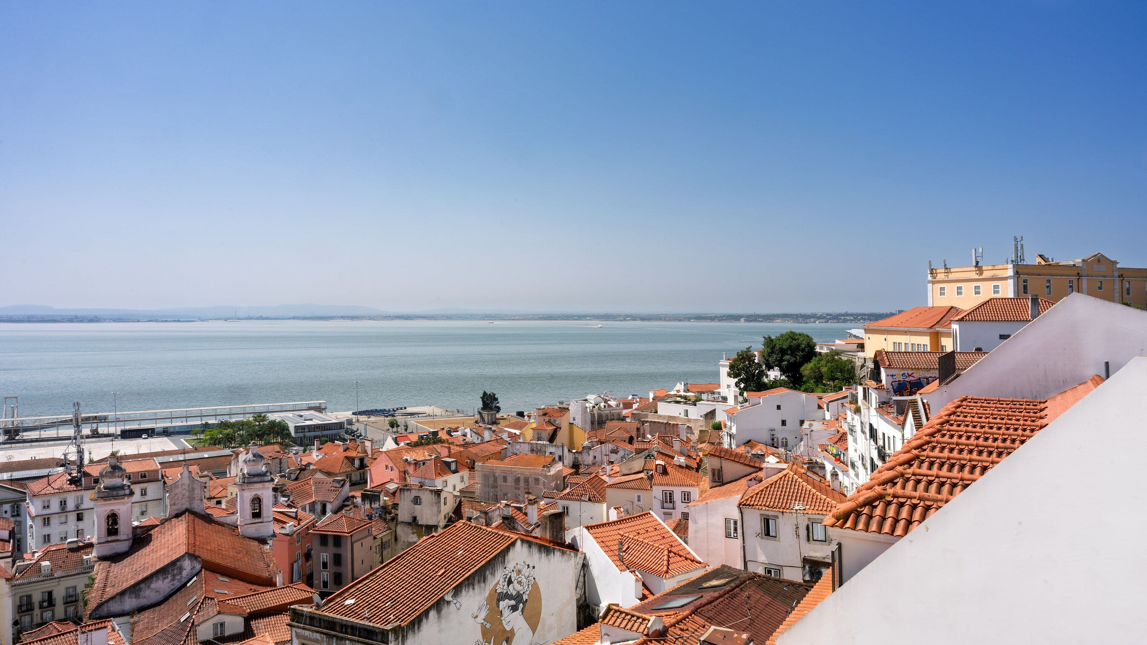 Tejados de teja roja del barrio de Alfama bajando hacia el estuario del Tajo, con la otra orilla al fondo