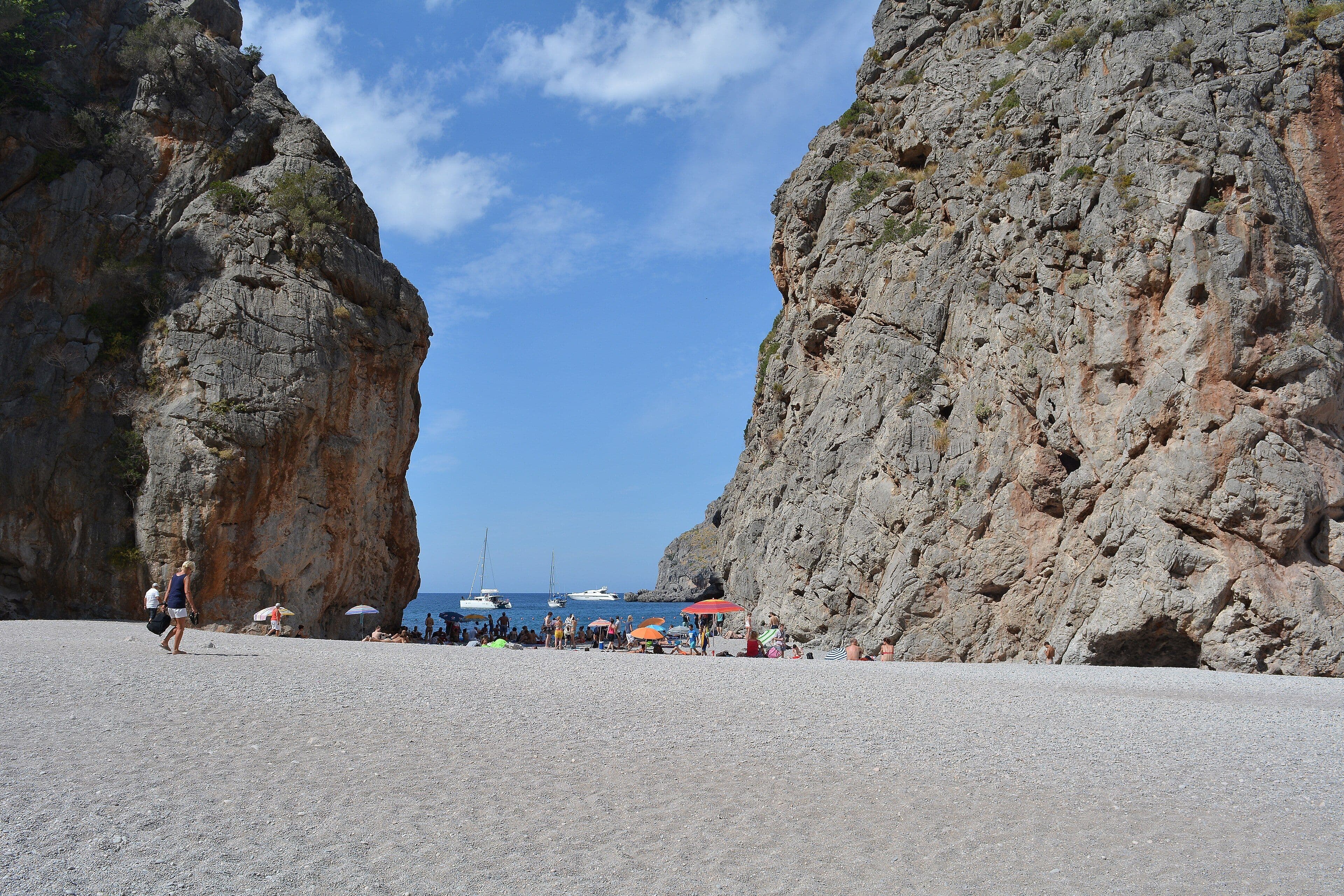 Playa de cantos blancos de Sa Calobra encajada entre dos grandes paredes de roca, con veleros fondeados en el mar al fondo